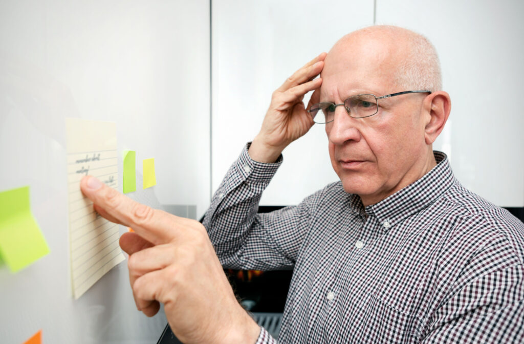 A senior with vascular dementia in a checkered long-sleeve shirt and glasses stands before a wall, gently touching his head as he struggles to recall the information written on the paper adhered to the wall