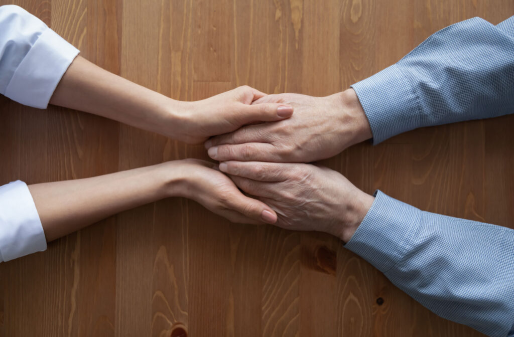 The hand of a female doctor holds onto a patient's hand as she engages in conversation with him.