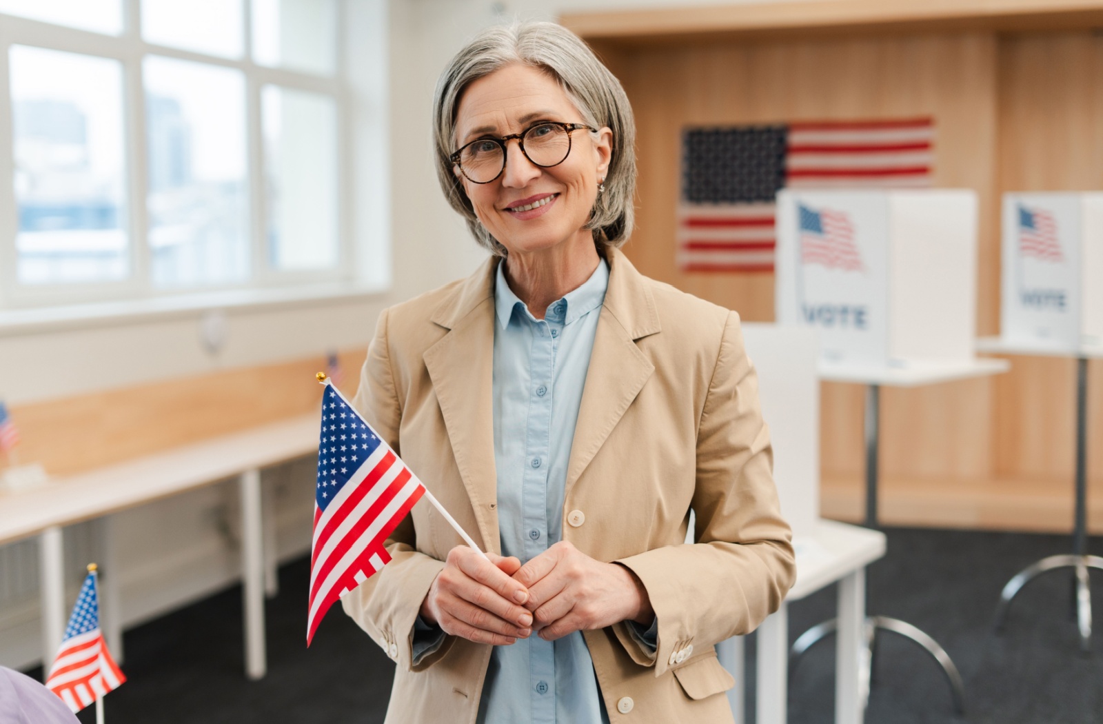 A smiling older adult with dementia holding a small American flag after voting in a public space.