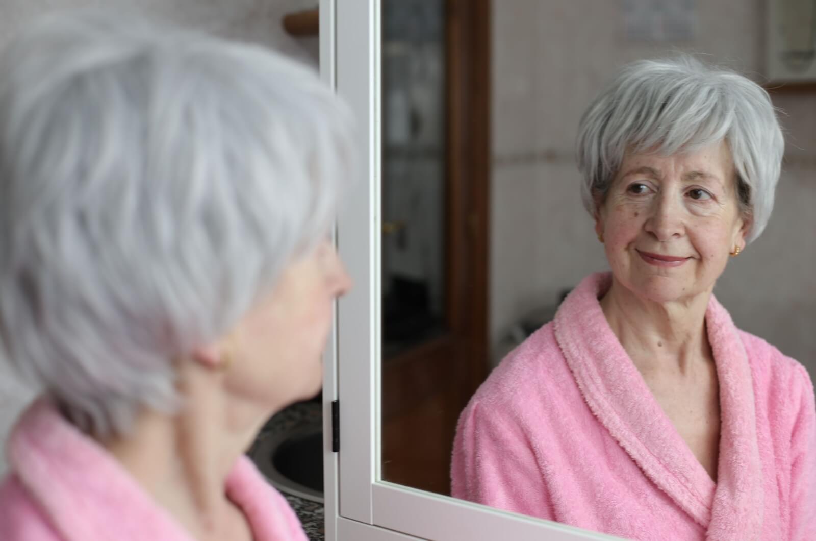 An older adult with dementia wearing a pink bathrobe and looking at their reflection in a bathroom mirror.