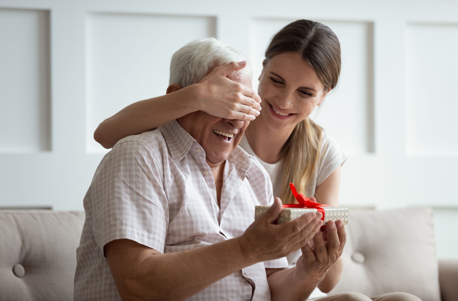 An adult covering the eyes of a parent with dementia while they laugh and prepare to open a meaningful gift.