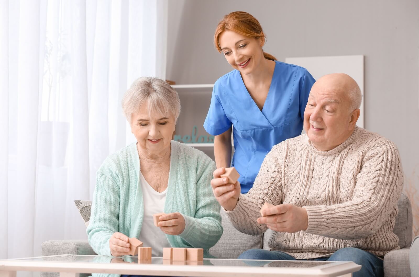 A smiling staff member in memory care standing behind two older adults sitting and holding wooden blocks.