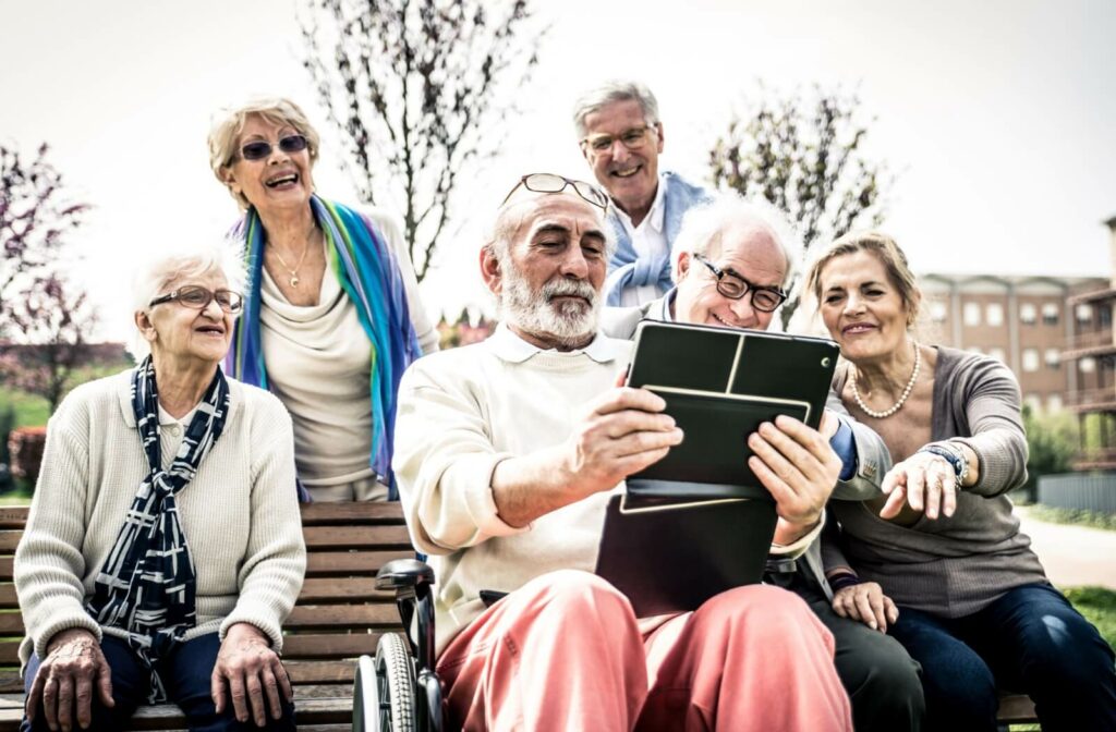 A smiling group of older adults looking at something funny on a tablet while outdoors.