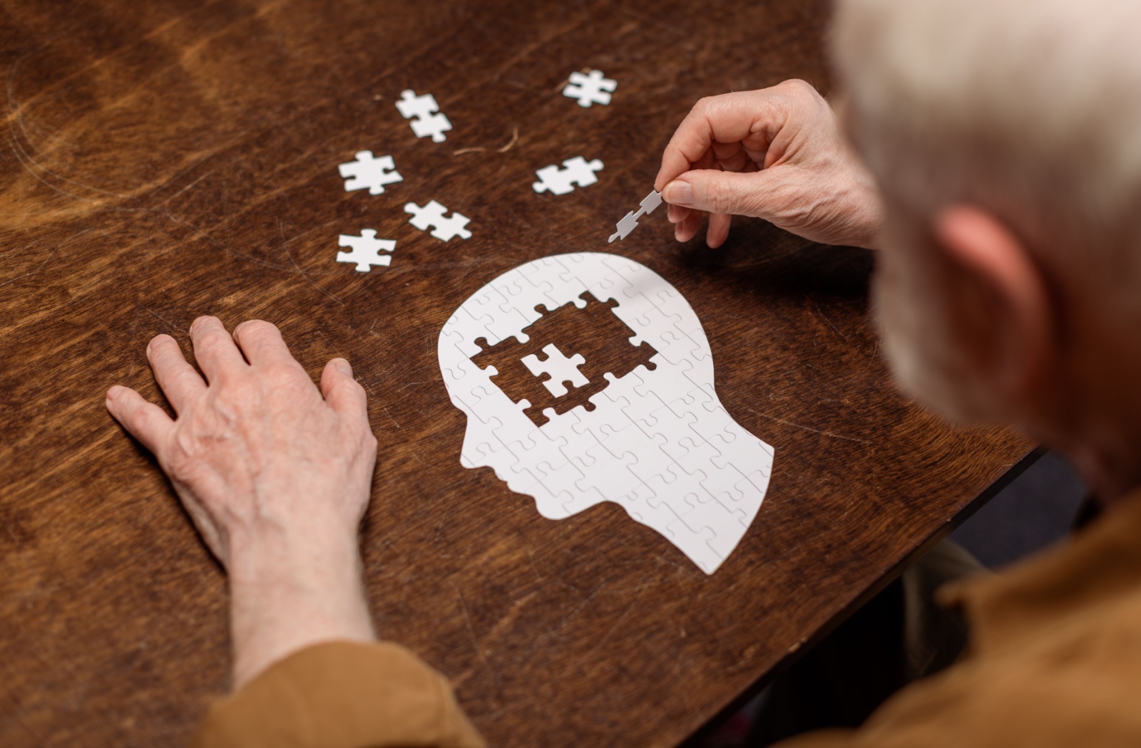 An over-the-shoulder view of an older man completing a puzzle shaped like the silhouette of a person's head.